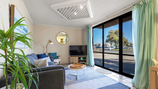 A modern living room with a sofa, a coffee table, a TV, and a large window with curtains. - Riverside Courtyard Glenelg North | Pets Welcome (Glenelg North)