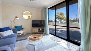 A living room with a sofa, TV, and a view of the street through large windows. - Riverside Courtyard Glenelg North | Pets Welcome (Glenelg North)