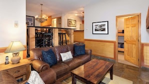 A living room with a leather sofa, wooden coffee table, and a chandelier. There is a staircase leading to a loft area.