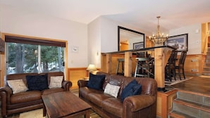 A living room with a brown leather sofa, two wooden coffee tables, a chandelier, and a staircase leading to an upper level.