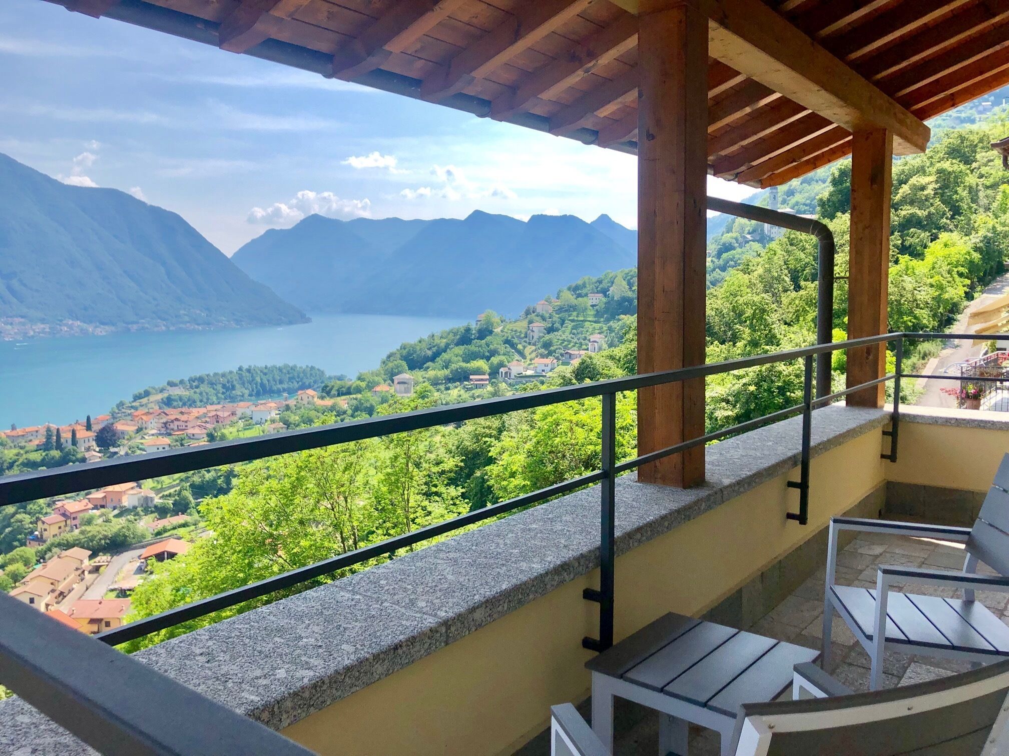 A balcony with a view of a lake and mountains, wooden furniture, and a stone balustrade.