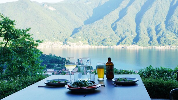 Una mesa con platos de comida, vasos y una botella de cerveza, con montañas y un lago al fondo.