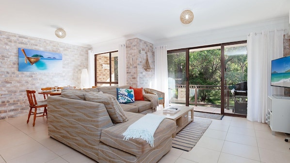 A spacious living room with a large sofa, a dining area, and a balcony with a view of greenery.