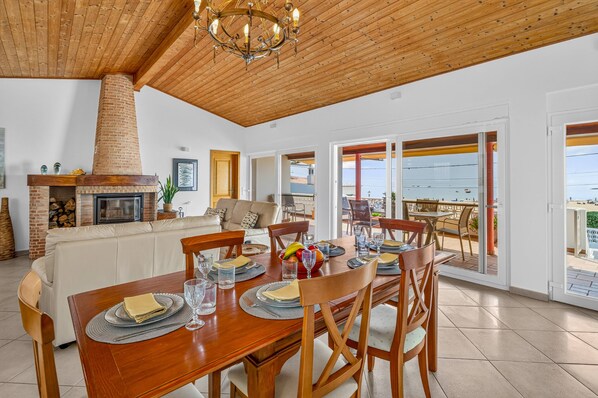 Un comedor con mesa de madera dispuesta para seis comensales, chimenea de ladrillo y vista al mar a través de puertas correderas amplias.