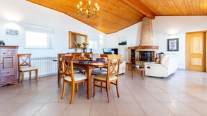 Een ruime woonkamer met houten plafond, een eettafel met stoelen, een open haard en een witte bank.