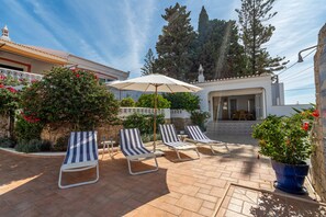 A patio with four striped lounge chairs, a white umbrella, and a stone wall with a window.