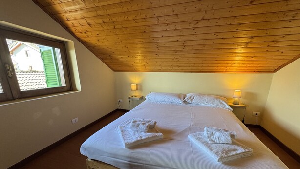 Une chambre à coucher avec un plafond en bois, un grand lit recouvert de draps blancs, deux lampes de chevet et une fenêtre avec des volets verts.