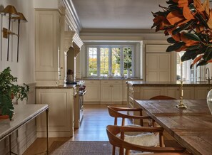 A kitchen with white cabinetry, a wooden dining table, and chairs. There is a window with a view of trees.