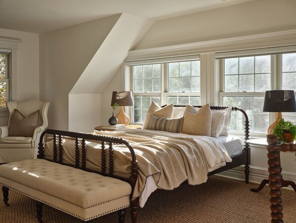 A bedroom with a large window, a bed with a wooden headboard, a bench, and a nightstand with a lamp.