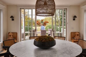 A round table with a bowl in the center, surrounded by wicker chairs, with a large wicker chandelier above.