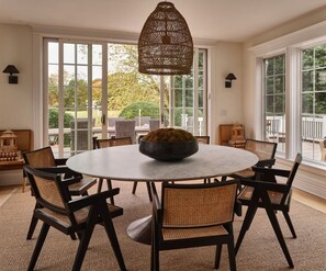 Une table de salle à manger ronde, surmontée d’un grand saladier, entourée de chaises en osier, dans une pièce dotée de grandes fenêtres et d’un luminaire suspendu.