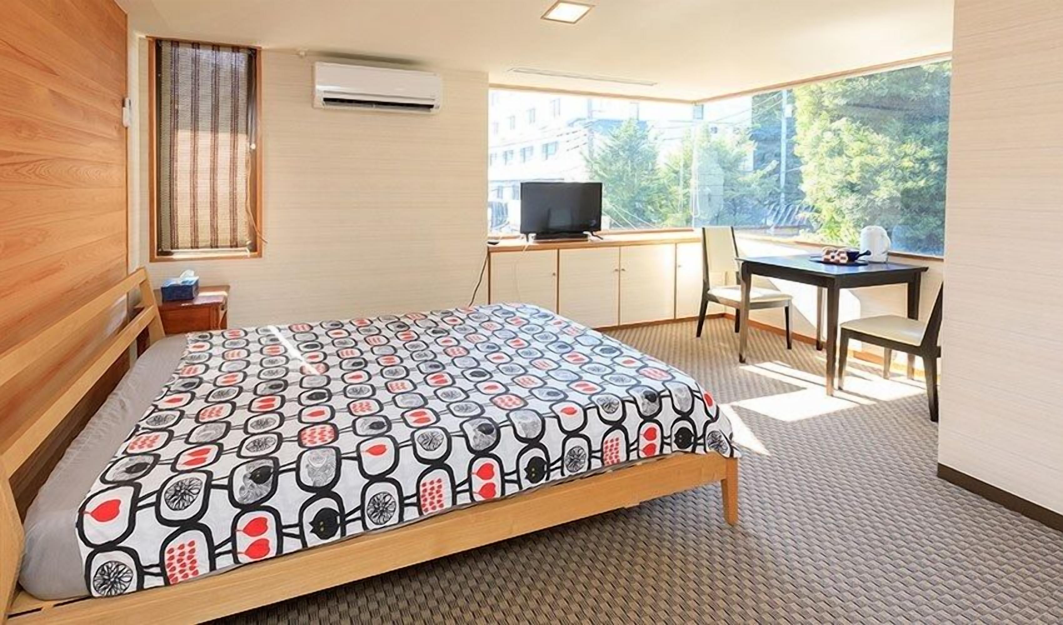 A bedroom with a bed, a desk, a chair, a television, and a window with a view of trees.