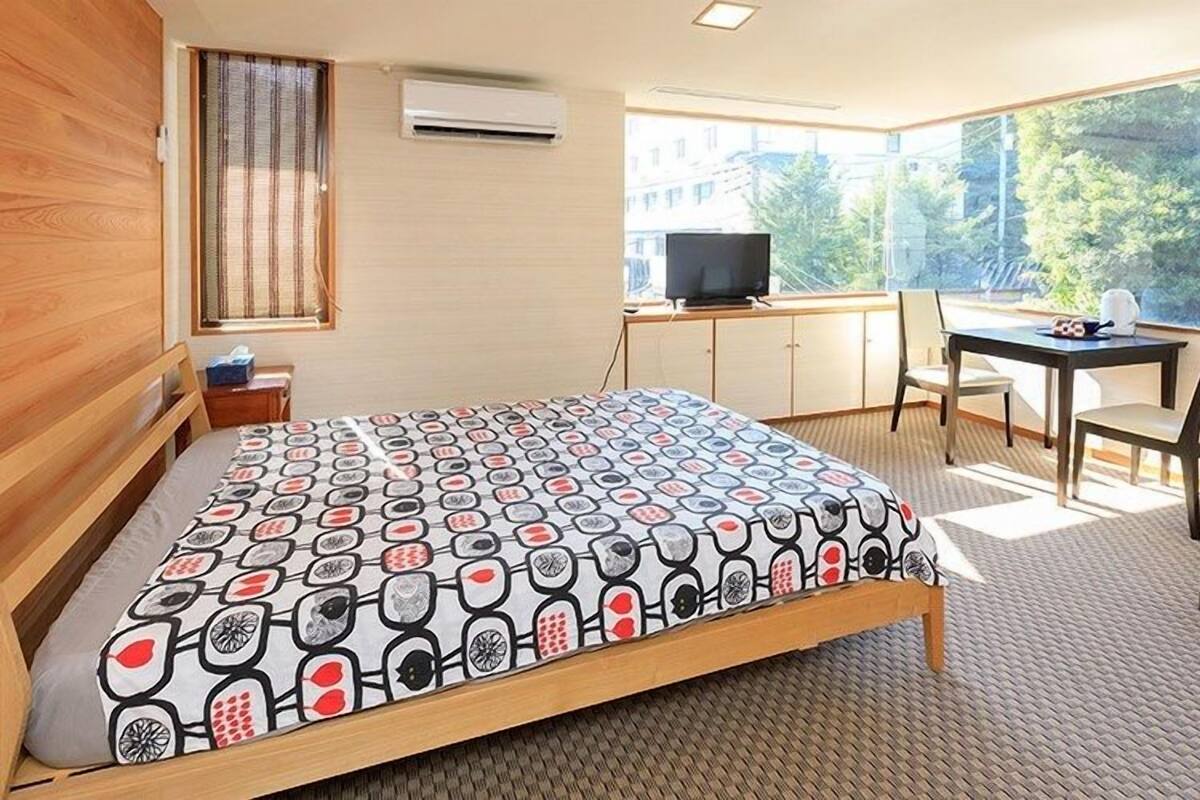 A bedroom with a bed, a desk, a chair, a television, and a window with a view of trees.