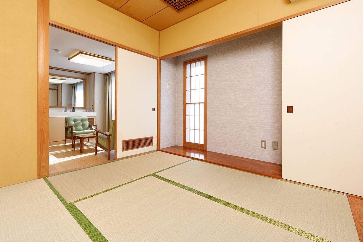 A traditional Japanese room with tatami flooring, sliding doors, and a view into another room with a green chair and a mirror.
