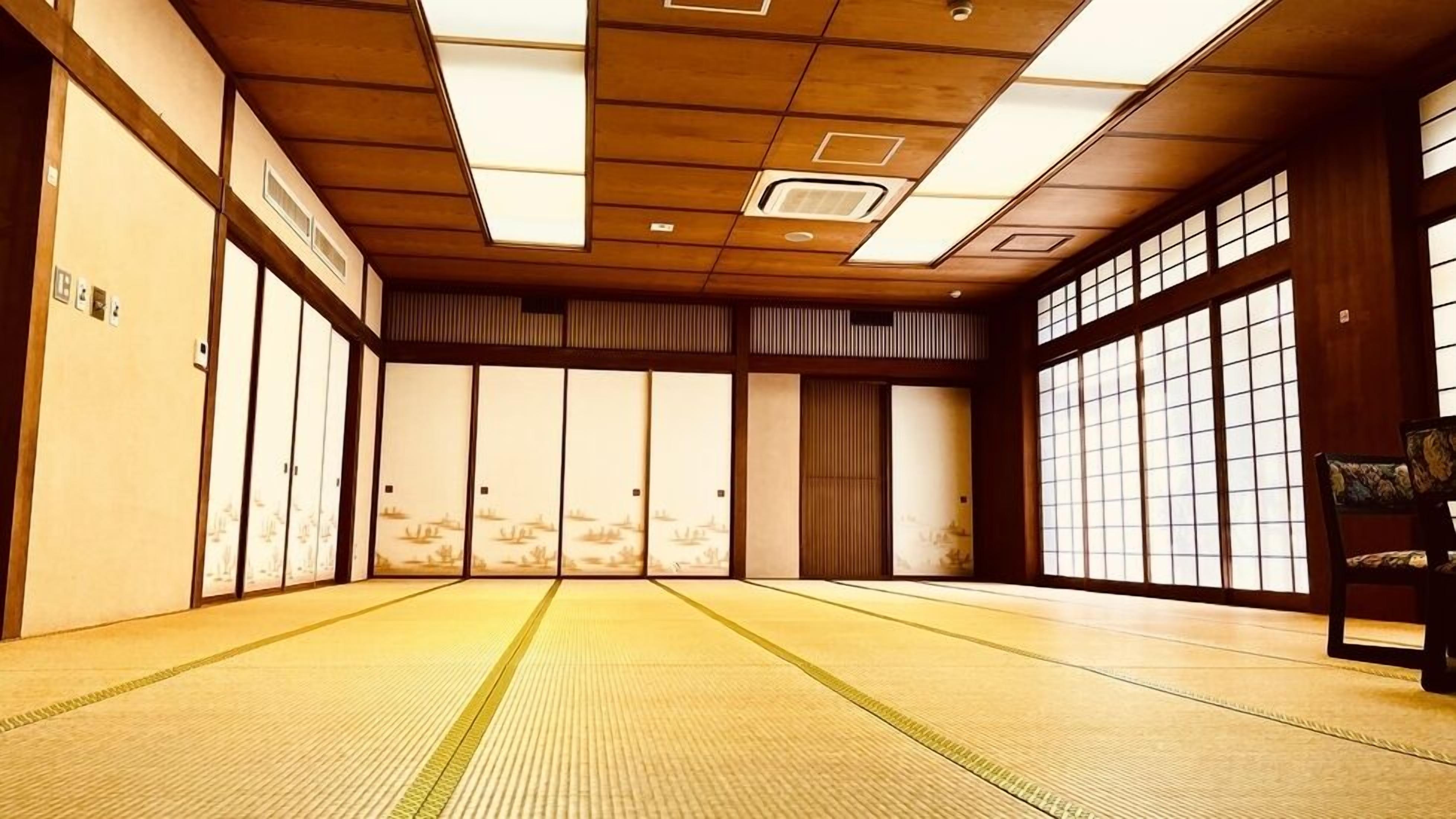 A traditional tatami mat room with sliding doors, a wooden ceiling with recessed lighting, and a view through the windows.