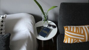 Una stanza con un cuscino bianco e nero, una poltrona scura con un cuscino giallo e bianco, e un tavolino rotondo con un libro, occhiali e una pianta.