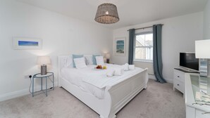Une chambre à coucher avec un lit blanc, une table de chevet avec une lampe, une télévision et une fenêtre avec des rideaux.