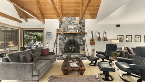 Un salon chaleureux avec une cheminée en pierre, des poutres apparentes en bois au plafond, un canapé gris, une table basse et un fauteuil en cuir noir.