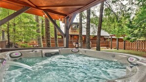 Un bain à remous avec une terrasse en bois et une vue sur un chalet en bois situé dans une zone boisée.