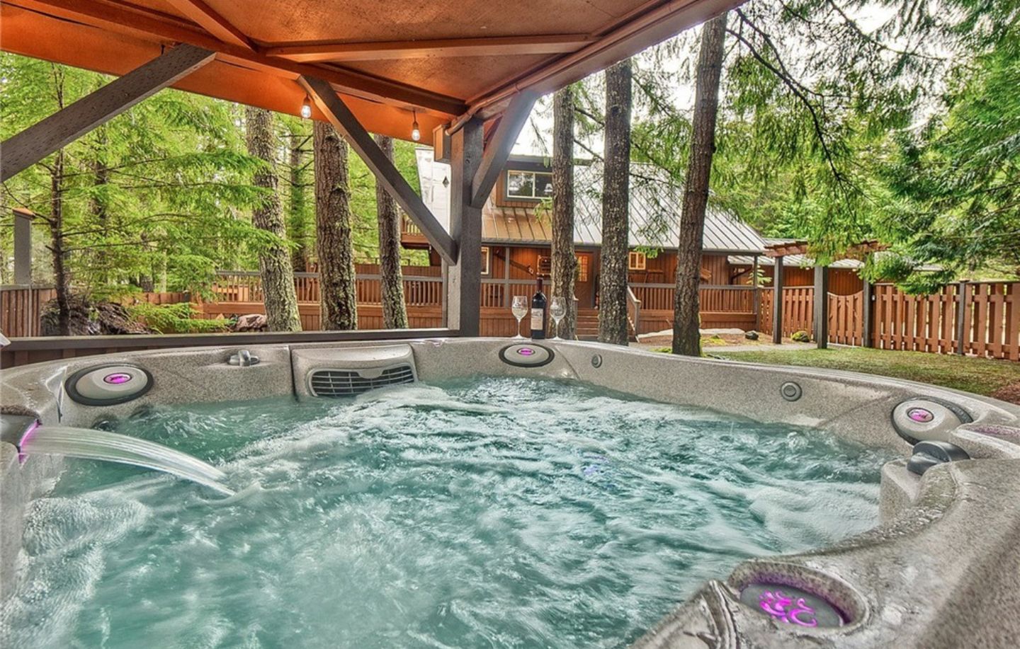 Ein Whirlpool mit Holzterrasse und Blick auf eine Holzhütte in bewaldeter Landschaft.
