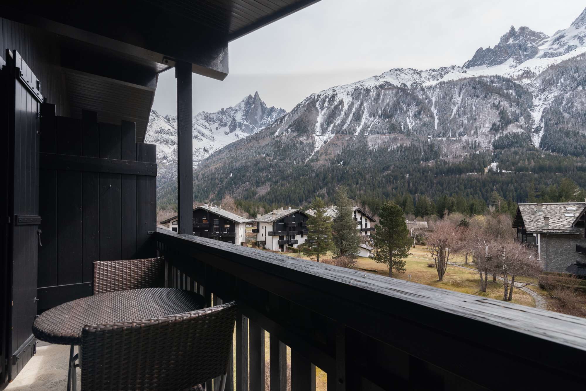 A balcony with wicker chairs overlooking a mountainous landscape with snow-capped peaks and a village below.