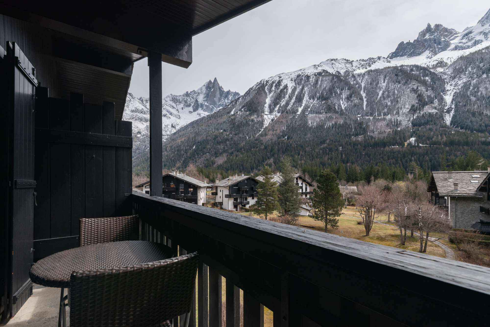 Un balcón con mobiliario de mimbre que ofrece vistas a un paisaje montañoso con cumbres nevadas y un pueblo abajo.
