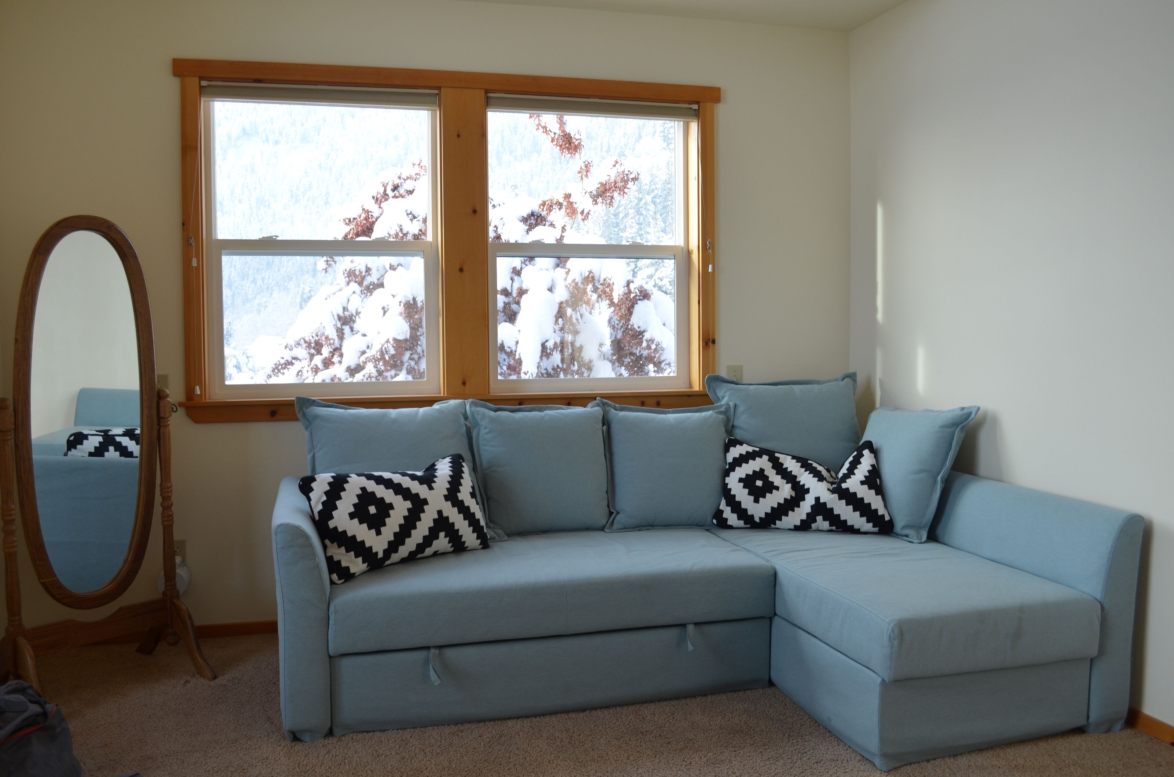 A light blue sectional sofa with patterned cushions, a wooden-framed window with a view of trees, and a full-length mirror.