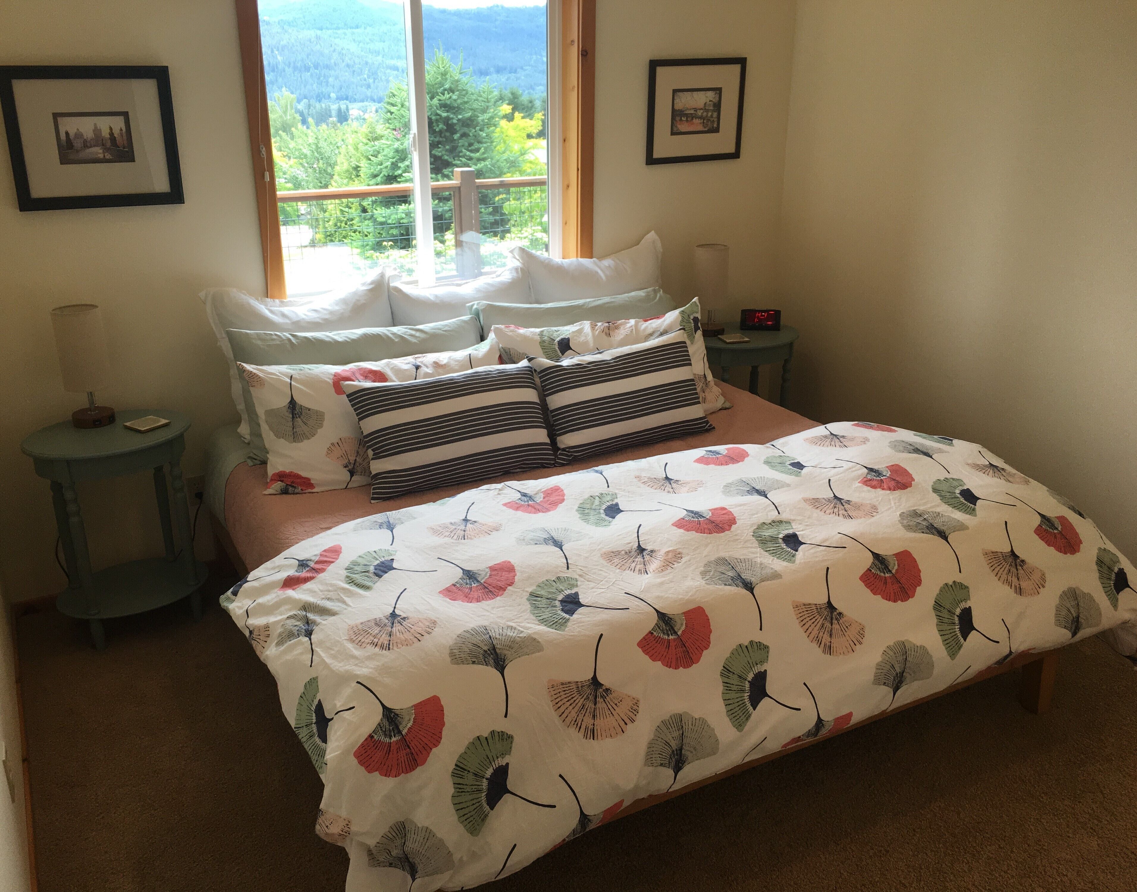 Quarto com cama, almofadas, mesa de cabeceira, abajur e quadros na parede.