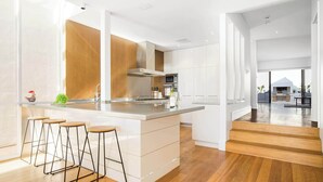 A modern kitchen with a central island, bar stools, and a view of the living area. - Spectacular Panoramic Oceanview House in Seacliff (Seacliff)