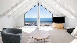 A high-ceiling room with a large bay window overlooking the sea, a flat-screen TV, and a wooden cabinet. - Spectacular Panoramic Oceanview House in Seacliff (Seacliff)