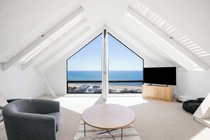 A high-ceiling room with a large bay window overlooking the sea, a flat-screen TV, and a wooden cabinet.