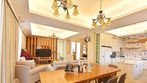 Uma sala de estar moderna com uma mesa de jantar de madeira, sofás e uma televisão. A cozinha possui armários brancos e painel de azulejos.