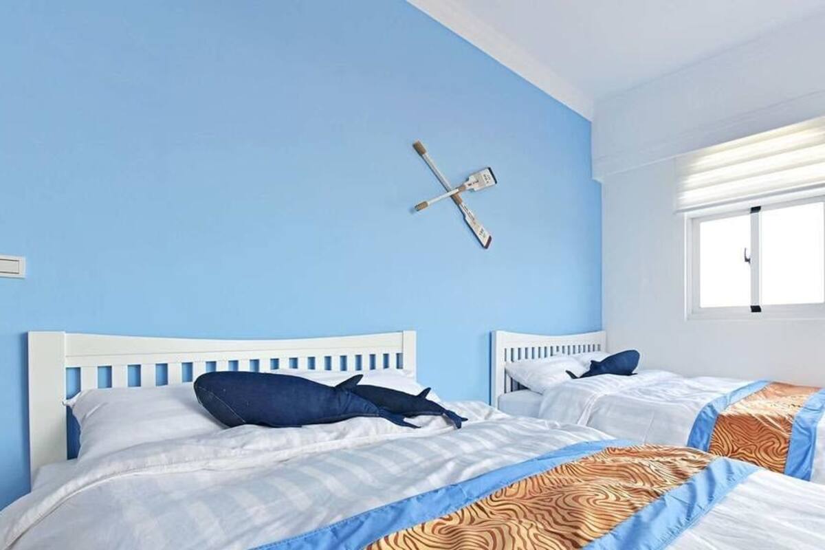 Una habitación con dos camas, cada una con cabecero blanco y ropa de cama azul. Se ve una ventana con persianas en la pared derecha, y una lámpara de pared sobre las camas.