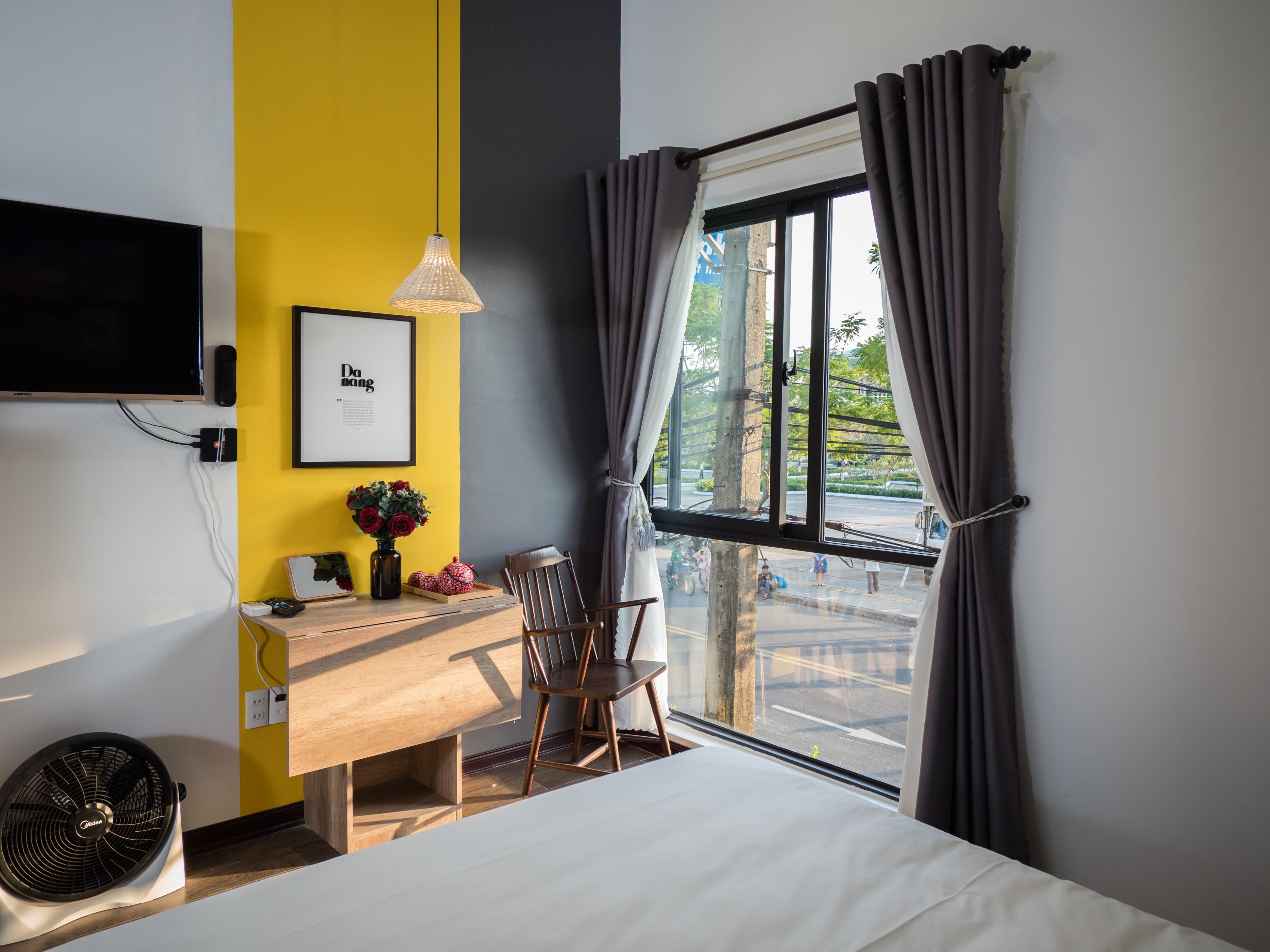 Une chambre d’hôtel comprenant un lit, un bureau avec une chaise, un téléviseur fixé au mur, une fenêtre avec des rideaux et un tableau encadré accroché au mur.