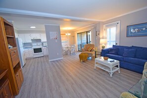 A modern living room with a blue sofa, a wicker coffee table, and a dining area with white chairs. - Our Place At The Beach 105E-Bayside 104th St, WIFI, Pool, W/D, AC (Ocean City)