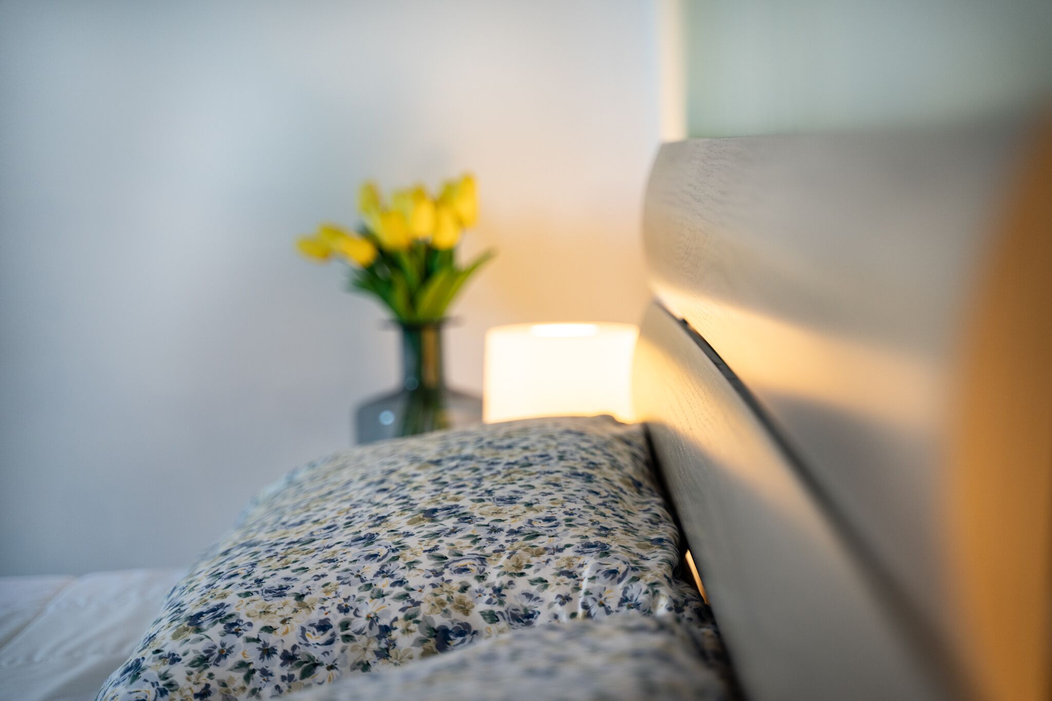 En seng med et blomstret sengetæppe, en læselampe og en vase med gule blomster på et natbord.