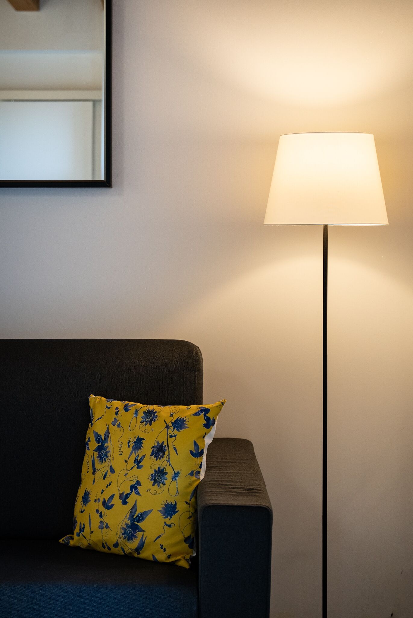 Un sofá de color oscuro con un cojín amarillo que presenta un estampado floral azul, ubicado al lado de una lámpara de pie con pantalla blanca.