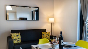 Uma sala de estar moderna com um sofá preto, almofadas florais amarelas, uma mesa de jantar posta com pratos, uma garrafa de vinho e um abajur.