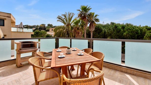 Una mesa de madera con sillas de mimbre en un balcón con vistas a un cuerpo de agua, con palmeras y edificios al fondo.