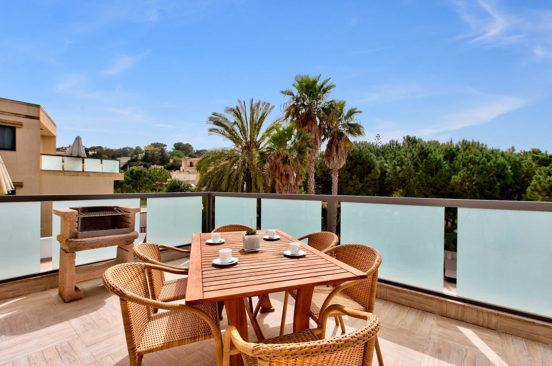 Een houten tafel met rieten stoelen op een balkon met uitzicht op water, met palmbomen en gebouwen op de achtergrond.