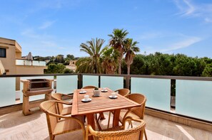 A wooden table with wicker chairs on a balcony overlooking a body of water, with palm trees and buildings in the background. - Villa Stephanotis~ Villa w/ Private Pool & Views (Il-Mellieħa)