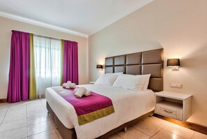 A hotel room with a large bed, a nightstand with a lamp, and a window with curtains. - Villa Stephanotis~ Villa w/ Private Pool & Views (Il-Mellieħa)