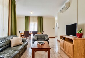 A living room with a leather sofa, a wooden coffee table, a TV on a wooden stand, and a dining area with a table and chairs. - Villa Stephanotis~ Villa w/ Private Pool & Views (Il-Mellieħa)