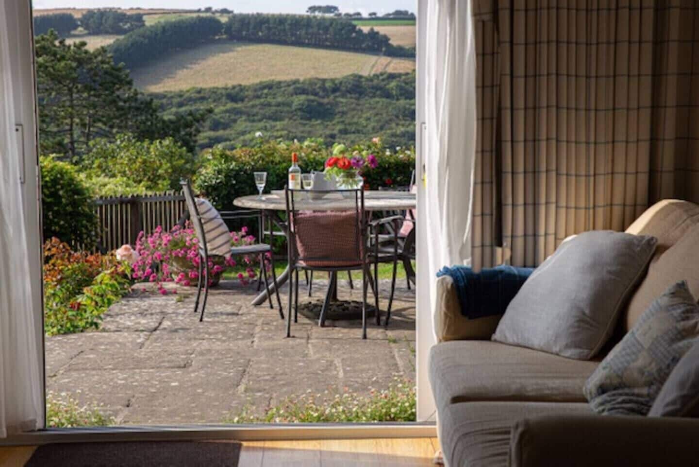 A room with a view of rolling hills, a dining area with a table and chairs, and a sofa with cushions.