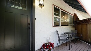 Une terrasse en bois avec une porte sombre, une fenêtre dotée d’un cadre blanc et une petite table avec des chaises.