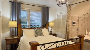 A bedroom with a wooden bed, two bedside tables with lamps, a window with blinds, a shower area, and a wardrobe. - 2 person jacuzzi suite with fireplace and kitchenette (Harrison Hot Springs)