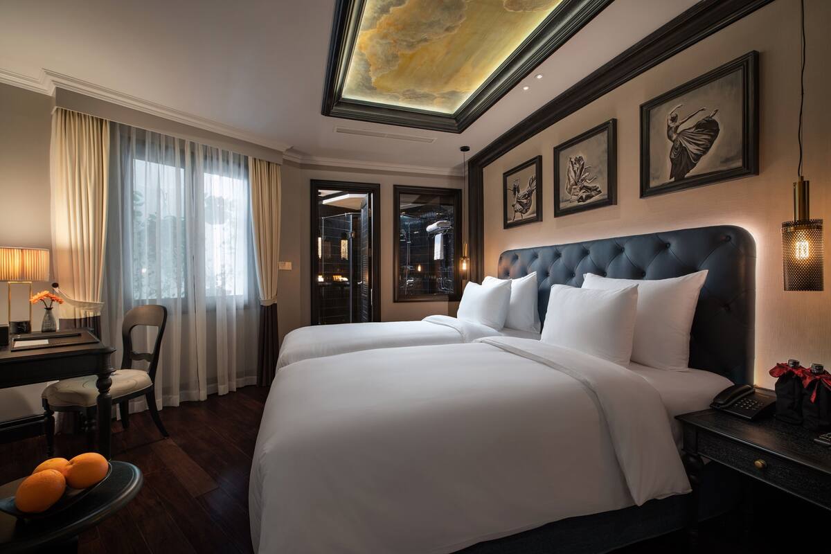 Una habitación de hotel con una cama grande, un escritorio con lámpara, una silla y un cuenco con naranjas.