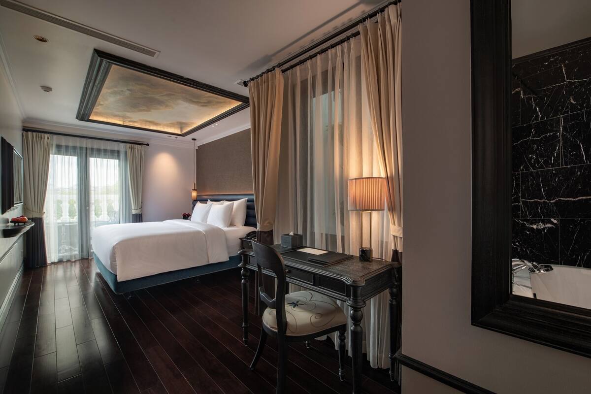 Habitación de hotel con una cama grande, un escritorio con silla, una lámpara y un baño con pared de mármol.