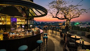 An outdoor bar area with a city view at sunset, featuring a bar counter, stools, and tables. - La Sinfonía del Rey Hotel & Spa (Hanoi)