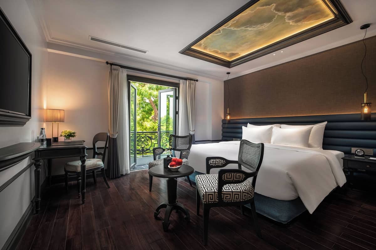 Habitación de hotel con una cama grande, un escritorio con silla, una mesita con frutero, y vistas a zonas verdes a través de un ventanal.